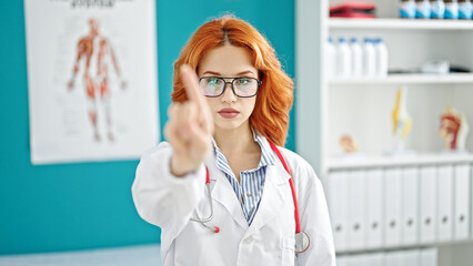 Young redhead woman doctor standing with serious expression saying no with finger at clinic