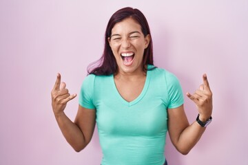 Young hispanic woman standing over pink background shouting with crazy expression doing rock symbol with hands up. music star. heavy music concept.