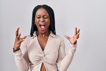 African woman with braids standing over white background crazy and mad shouting and yelling with...