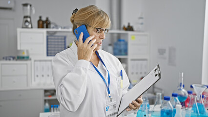Blonde middle age female scientist engrossed in a serious talk over the smartphone while reading a...