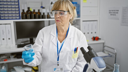 Dedicated middle age blonde woman scientist, safely clad in glasses and gloves, meticulously measures test tube contents in a bustling lab.
