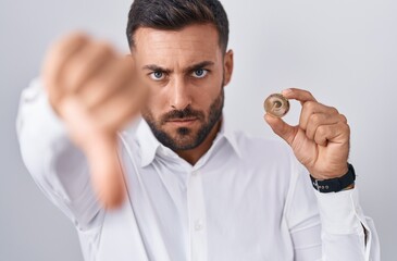 Handsome hispanic man holding polkadot cryptocurrency coin with angry face, negative sign showing...