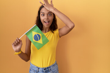 Young hispanic woman holding brazil flag surprised with hand on head for mistake, remember error....