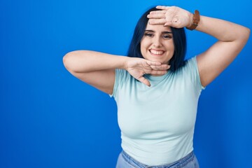 Obraz na płótnie Canvas Young modern girl with blue hair standing over blue background smiling cheerful playing peek a boo with hands showing face. surprised and exited