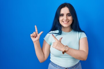 Fototapeta premium Young modern girl with blue hair standing over blue background smiling and looking at the camera pointing with two hands and fingers to the side.