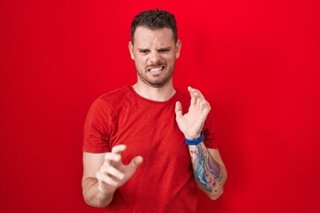 Young hispanic man standing over red background disgusted expression, displeased and fearful doing disgust face because aversion reaction.