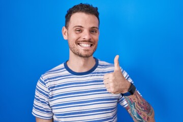 Fototapeta premium Young hispanic man standing over blue background doing happy thumbs up gesture with hand. approving expression looking at the camera showing success.