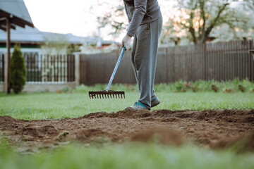 Adult man in gray tracksuit standing on green lawn and working soil with garden rake in hands near...