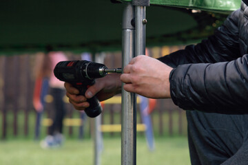 Close up of hands of man fastening metal frame stand to trampoline support using cordless...