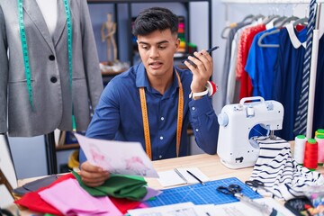 Young hispanic man tailor talking on smartphone looking clothing design at clothing factory