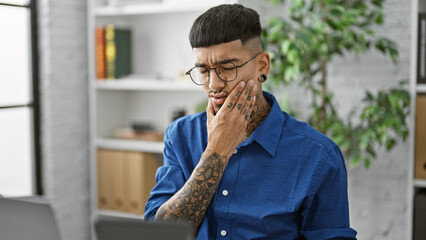 Struggling at work, young, handsome latin business worker fights through toothache pain while seriously working in his office interior