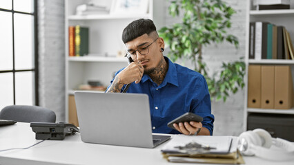 Young, serious latin man engrossed in conversation, talking on the phone while working on his laptop at the office. busy professional, overworked employee or tattooed boss managing task