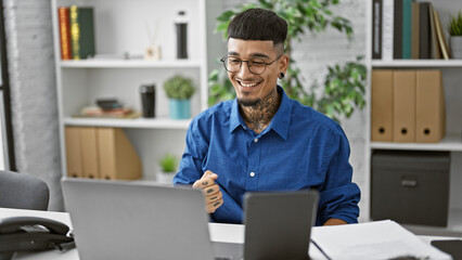 Triumphant latin young man sitting in the office, working on laptop and touchpad, celebrating business success with a charismatic smile, embodying the winning spirit of a dedicated professional.