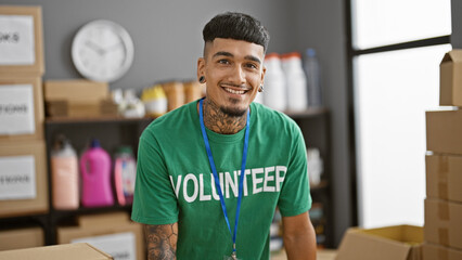 Confident young latin volunteer man, smiling and standing in his t-shirt at the heart of charity center, enjoying his altruistic work within the warm community