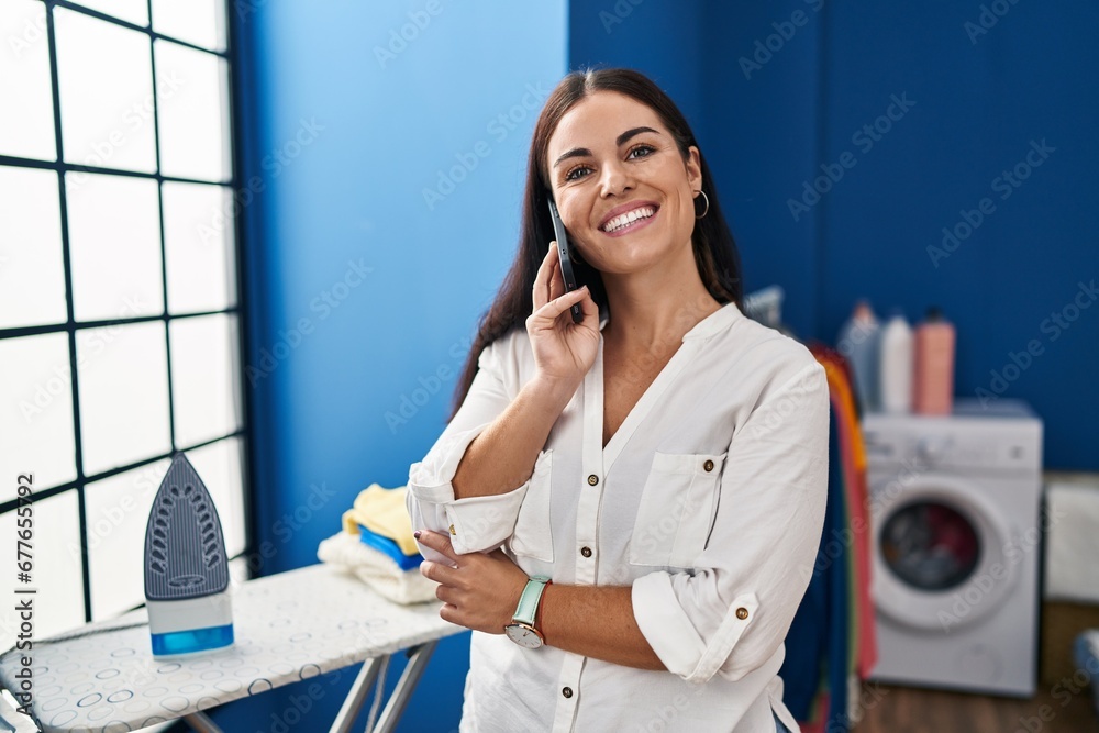 Sticker Young beautiful hispanic woman talking on smartphone standing at laundry room