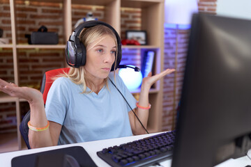 Young caucasian woman playing video games wearing headphones clueless and confused expression with...