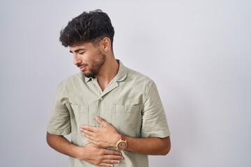 Arab man with beard standing over white background with hand on stomach because indigestion,...