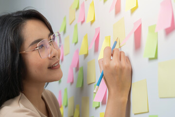 Happy Asian business woman write short note to do list in sticker on wall in meeting office room.