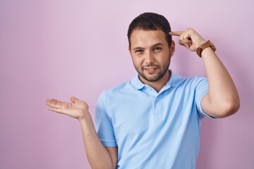 Fototapeta premium Hispanic man standing over pink background confused and annoyed with open palm showing copy space and pointing finger to forehead. think about it.