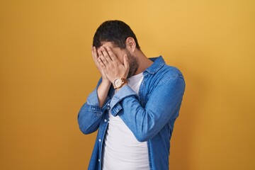 Hispanic man standing over yellow background with sad expression covering face with hands while...
