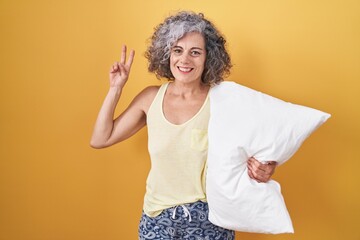 Middle age woman with grey hair wearing pijama hugging pillow smiling looking to the camera showing...