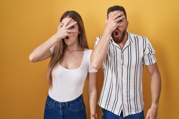 Young couple standing over yellow background peeking in shock covering face and eyes with hand, looking through fingers with embarrassed expression.