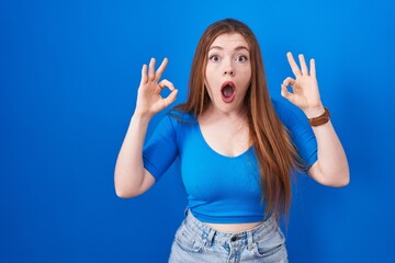 Redhead woman standing over blue background looking surprised and shocked doing ok approval symbol with fingers. crazy expression