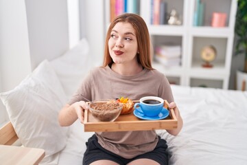 Redhead woman wearing pajama holding breakfast tray smiling looking to the side and staring away...