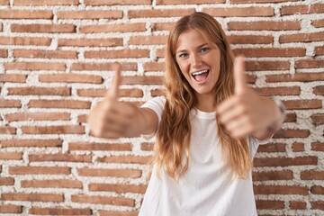 Young caucasian woman standing over bricks wall approving doing positive gesture with hand, thumbs up smiling and happy for success. winner gesture.