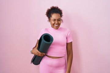 Young hispanic woman with curly hair holding yoga mat over pink background winking looking at the...
