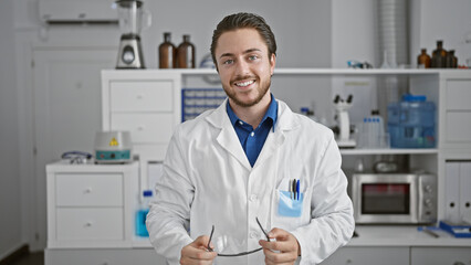 Young hispanic man scientist holding glasses smiling at laboratory