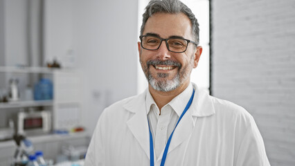 Brimming with confidence, smiling young hispanic man, grey-haired scientist standing in the lab amidst experiments