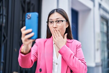 Chinese young woman doing video call with smartphone covering mouth with hand, shocked and afraid...