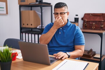 Young hispanic man working at the office with laptop smelling something stinky and disgusting, intolerable smell, holding breath with fingers on nose. bad smell - obrazy, fototapety, plakaty