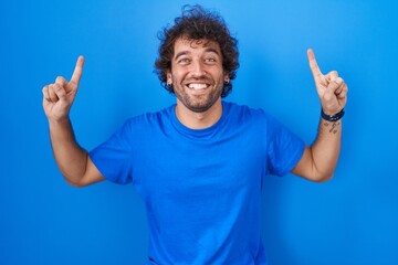 Naklejka premium Hispanic young man standing over blue background smiling amazed and surprised and pointing up with fingers and raised arms.