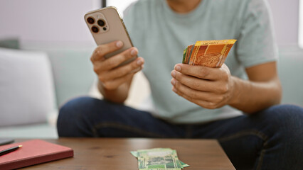 Young hispanic man chilling on sofa, happily counting his stash of south african rand banknotes, busy texting about his flourishing business from his cosy living room - obrazy, fototapety, plakaty