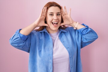 Young hispanic woman with red hair standing over pink background smiling cheerful playing peek a boo with hands showing face. surprised and exited