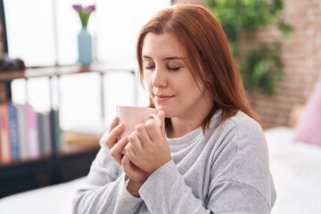 Young beautiful plus size woman smelling cup of coffee sitting on bed at bedroom