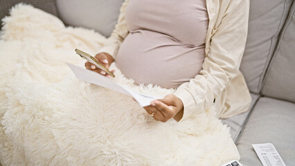 Tucked in a blanket at home, a young, pregnant woman, warmly seated on her sofa, indulges in...