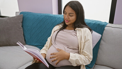 Glowing young pregnant woman enjoying comfortable rest, engrossed in reading book while touching...