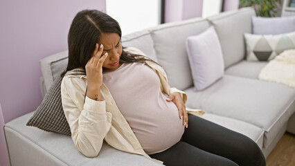 Young hispanic woman suffering a serious headache during pregnancy, sitting at home and massaging belly stressfully