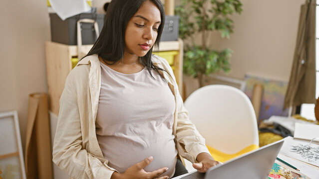 Eager young pregnant woman sitting at her art studio table mastering her craft, painting on canvas with laptop as guide.