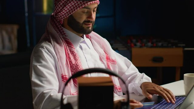 Smilling arab man doing online shopping, using computer to look up tech products to buy. Middle Eastern person adding credit card payment method on secure website, ordering with instant delivery