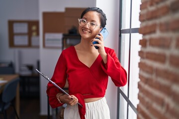 Young chinese woman business worker talking on smartphone holding clipboard at office