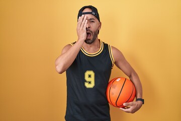 Middle age bald man holding basketball ball over yellow background yawning tired covering half face, eye and mouth with hand. face hurts in pain.