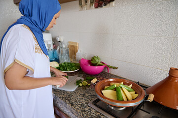 Muslim housewife woman chopping green beans on cutting board, cooking Moroccan tagine standing by marble kitchen counter