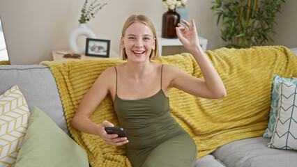 Young blonde woman using smartphone doing ok gesture at home