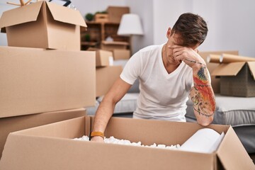 Young man unpacking cardboard box with unhappy expression at new home - obrazy, fototapety, plakaty