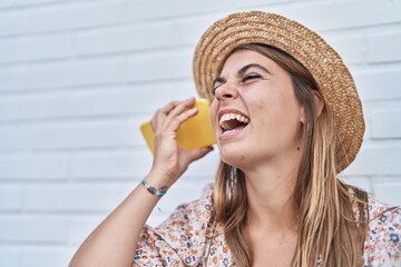 Young woman tourist wearing summer hat listening voice message by smartphone over isolated white brick background