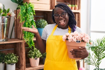 Young african woman working at florist shop holding plant celebrating achievement with happy smile...